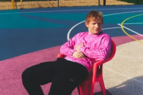 Ed Sheeran Konzert 2025 München Ticket-Verkauf - hier ein Pressefoto von Ed Sheeran 2025. bei dem der Künstler auf einem Stuhl in einem Stadion bzw. auf einem Sportplatz sitzt