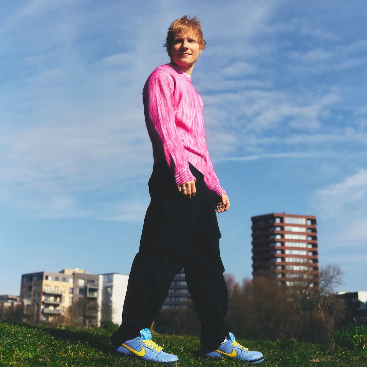 Ed Sheeran - neues Album „Play“ ein musikalisches Highlight - hier ein Pressefoto von Ed Sheeran 2025, bei dem der Künstler auf einer Wiese steht mit einer typischen Wohnstadt im Hintergrund