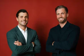 Die zwei Bachelors 2026 Tim Reitz und Sebastian Paul. Das Foto zeigt die beiden Männer in einem Fotostudio-Ambiente vor einer weinroten Wand oder Fläche, sie halten die Arme vor der Brust verschränkt und lächeln in die Kamera.