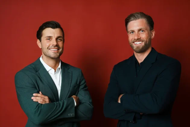 Die zwei Bachelors 2026 Tim Reitz und Sebastian Paul. Das Foto zeigt die beiden Männer in einem Fotostudio-Ambiente vor einer weinroten Wand oder Fläche, sie halten die Arme vor der Brust verschränkt und lächeln in die Kamera.