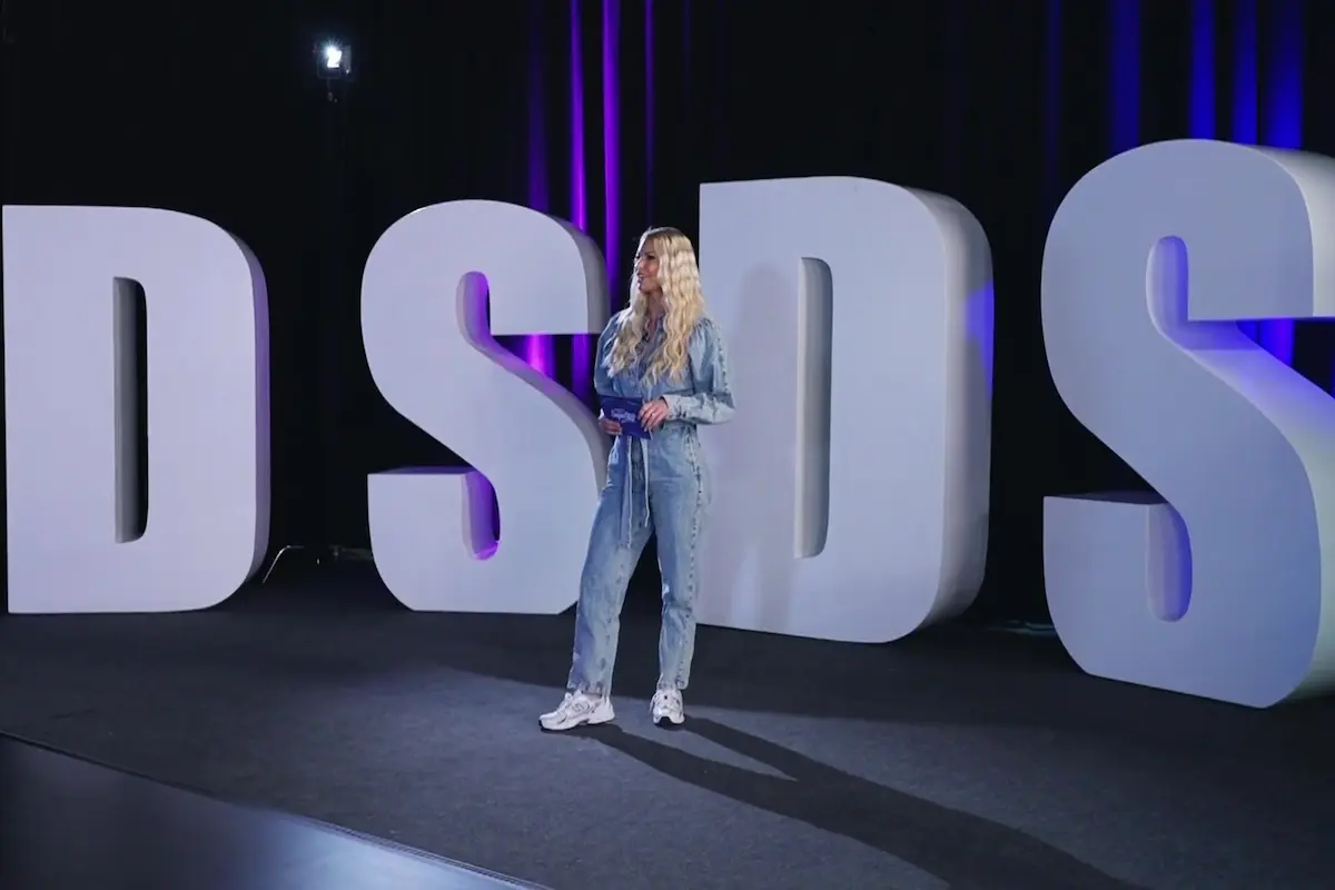 DSDS 2027 - Übersicht - hier im Bild die Buchstaben DSDS in der Größe eines Menschen - davor steht Isi Glück (in einem Jeans-Overall mit offenen Haaren), die wieder Mitglied in der Jury bei DSDS 2027 sein wird