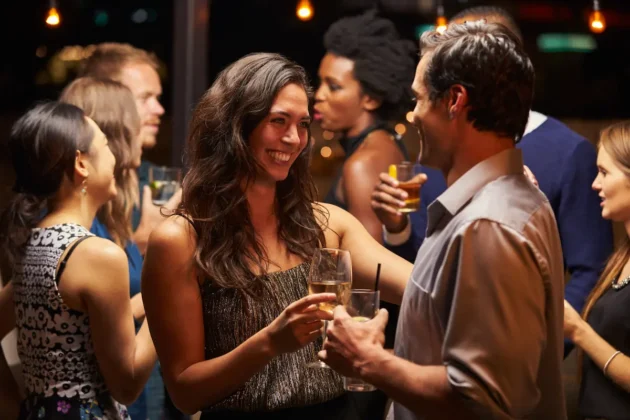 Ist ein Schnuppertanzkurs beim ersten Date eine gute Idee? Ja! - hier im Bild zu sehen ist ein Pärchen in einer Bar, das sich anlächelt - direkt hinter dem Pärchen steht eine Gruppe weiterer Gäste, die sich unterhalten