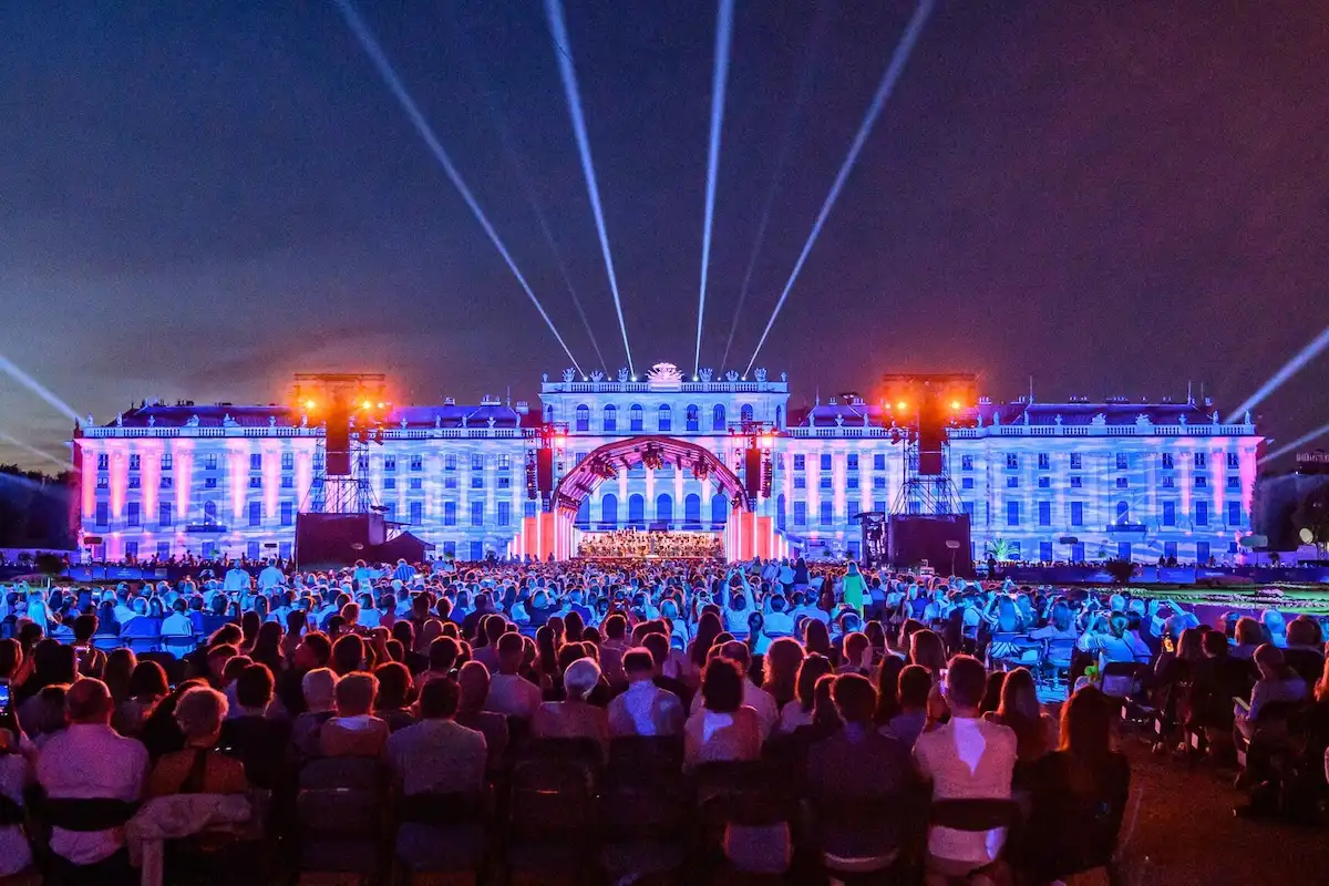 Sommernachtskonzert 2026 Wiener Philharmoniker 19.6.2026 in Wien - hier im Bild die Bühne mit dem Orchester vor dem stimmungsvoll angeleuteten Schloss Schönbrunn, im Vordergrund sitzen Zuschauer